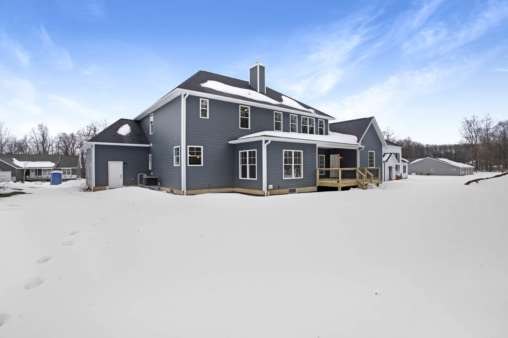 Two-story house with wooden deck and black railing, snow covering roof and large yard, white door with black knobs, white-framed windows, bare trees in winter landscape