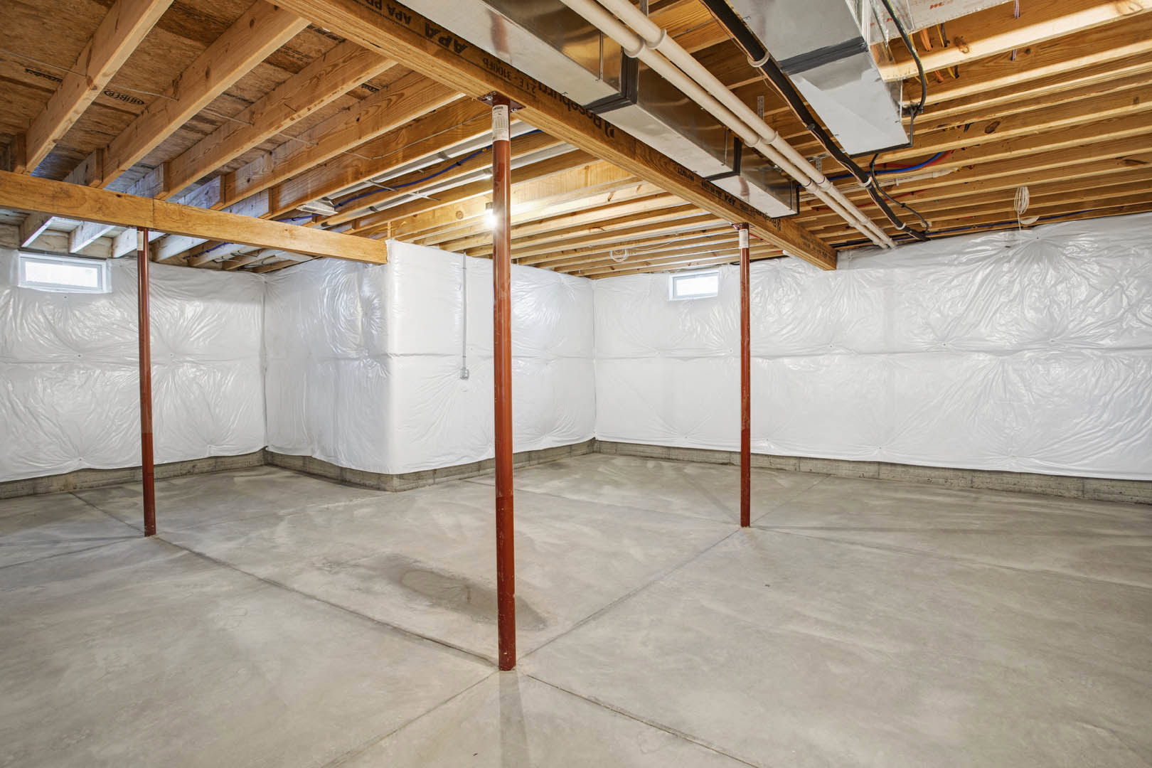 Basement with white plaster walls, exposed wooden ceiling beams, concrete floor, brown support pole, and white plastic insulation covering