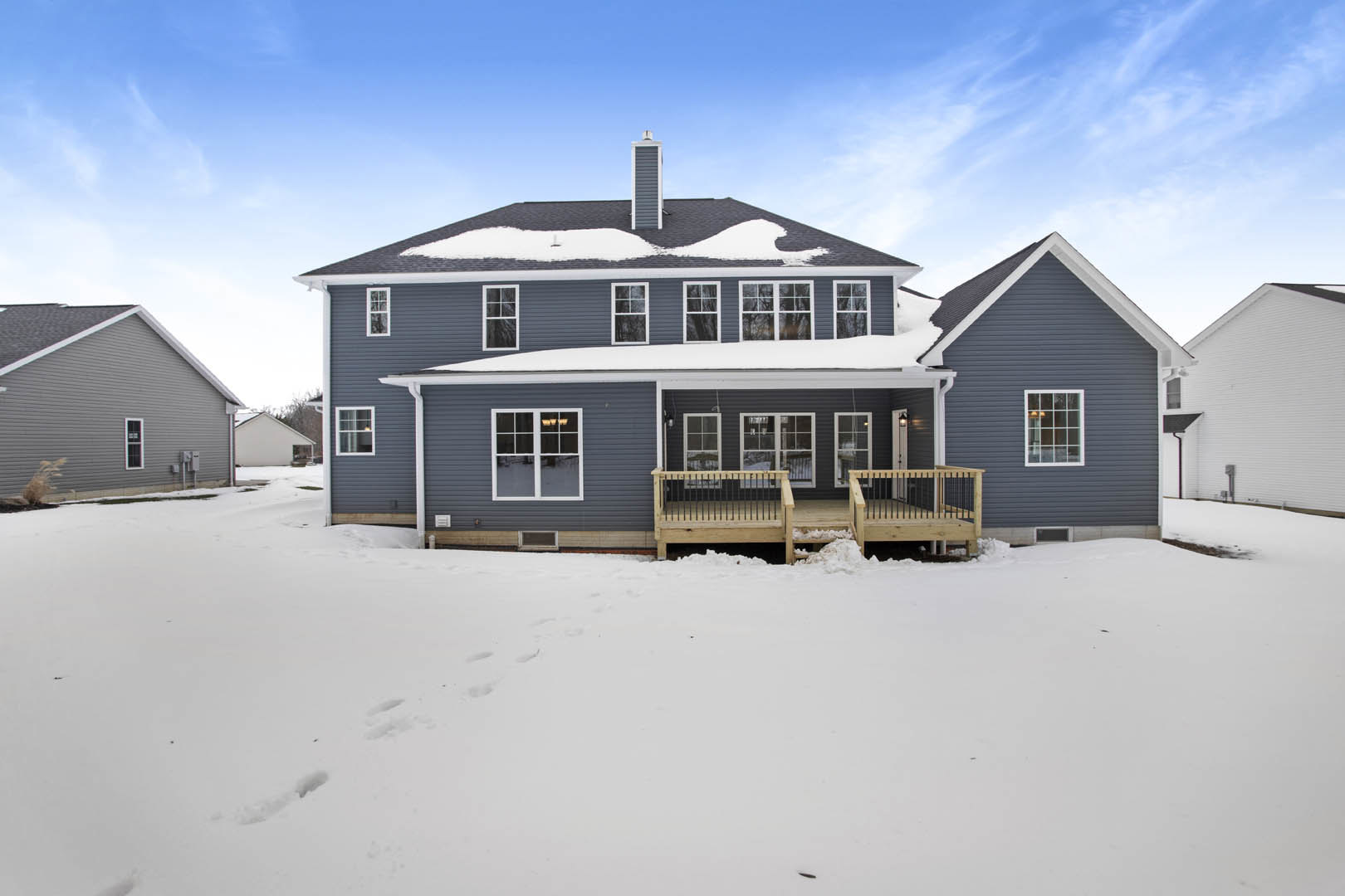 Grey siding house with white roof, wooden deck partially covered in snow, multi-pane window visible, winter landscape with snow on ground and roof