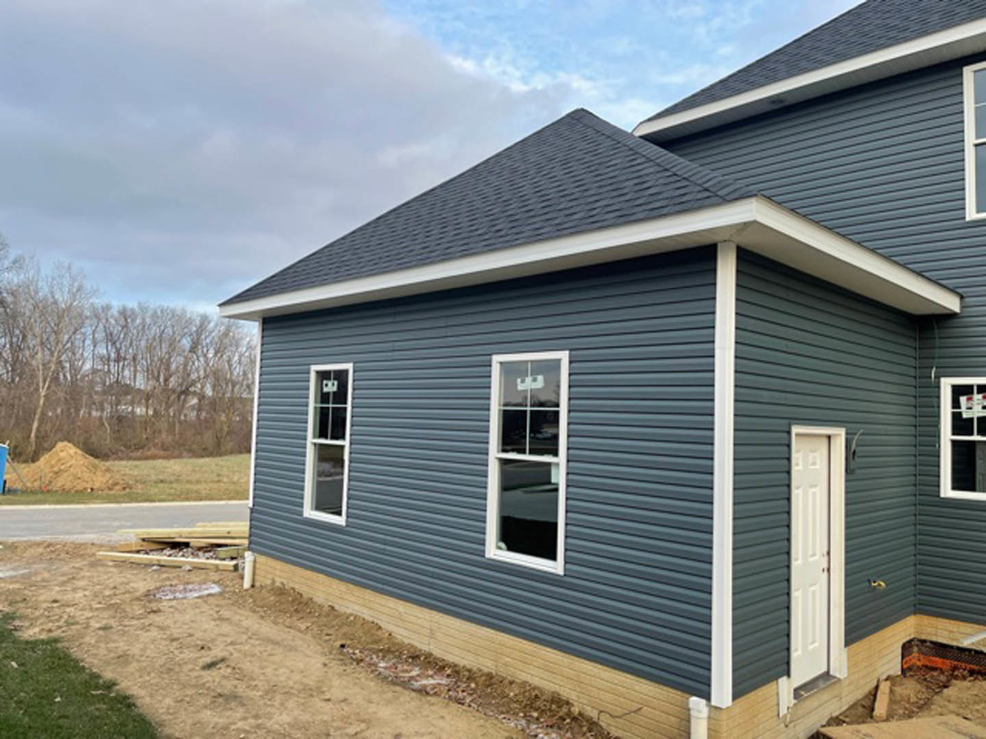 Partially built house with exposed framing, white door and window, dirt path leading to entrance, piles of wood and rocks in foreground, surrounded by mature trees under a cloudy