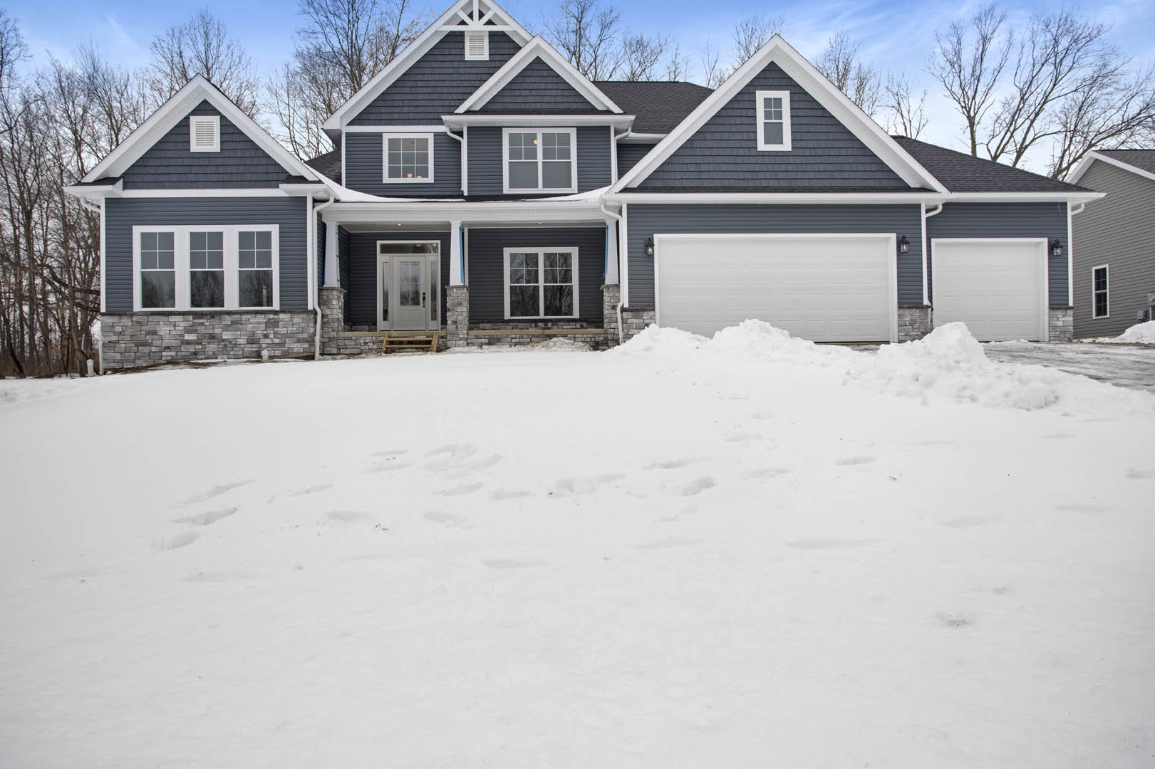 Two-story house with white siding, row of windows, and attached garage; snow covers the ground with visible footprints, trees in background.