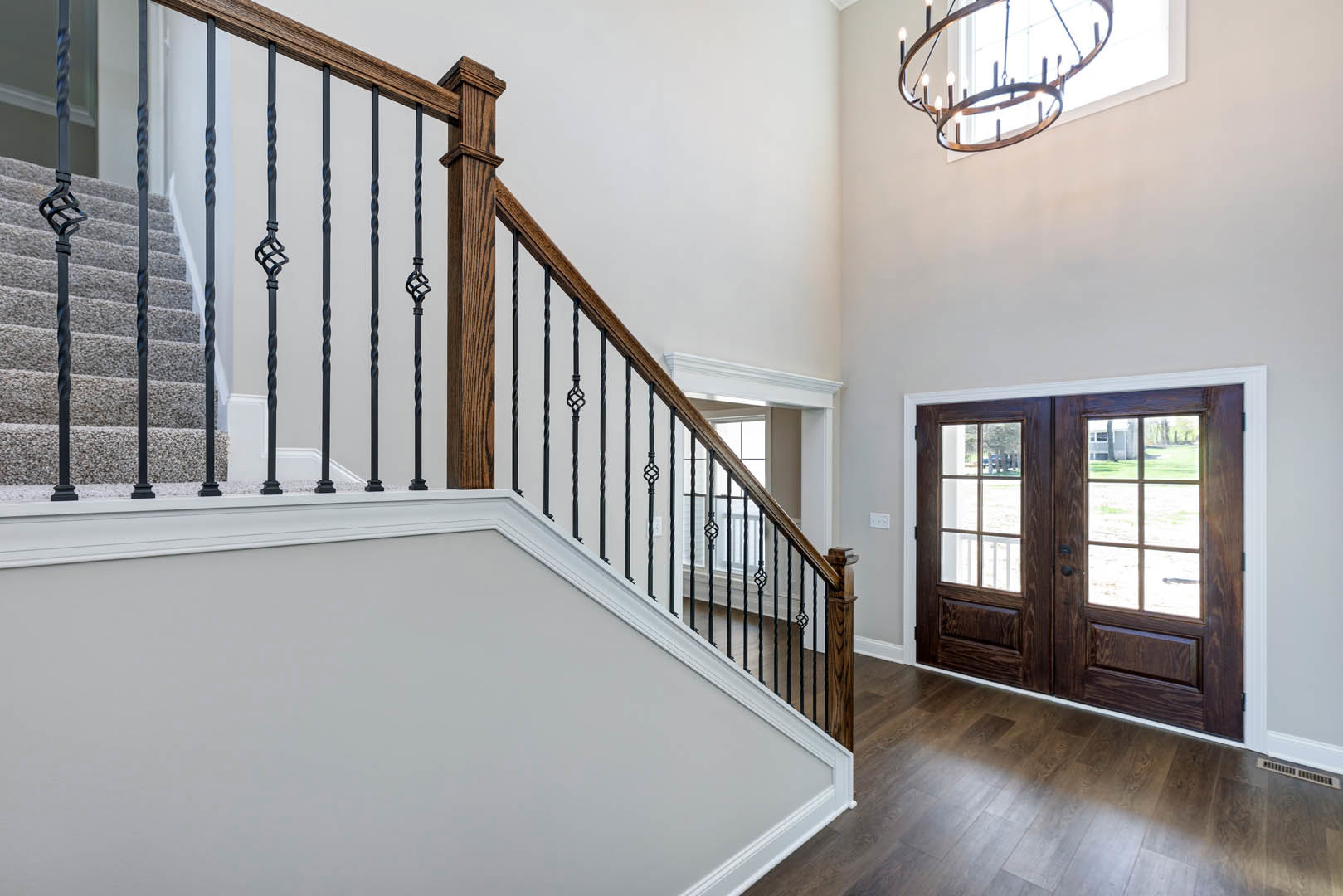 Curved staircase with black metal railing, wooden steps, glass-paneled double doors, chandelier hanging from ceiling, light wood flooring, white walls with crown molding