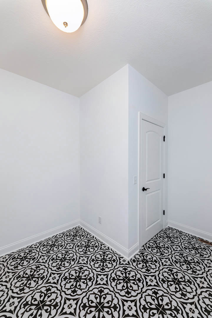 White door with black handle set in a white room featuring black and white patterned tile flooring, white plaster walls, and a ceiling light fixture.