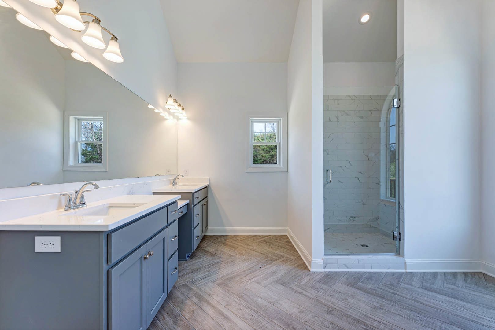 Bathroom with glass shower enclosure, white tile walls, double sinks on white countertop, window overlooking trees, modern light fixture, and white electrical outlet with black