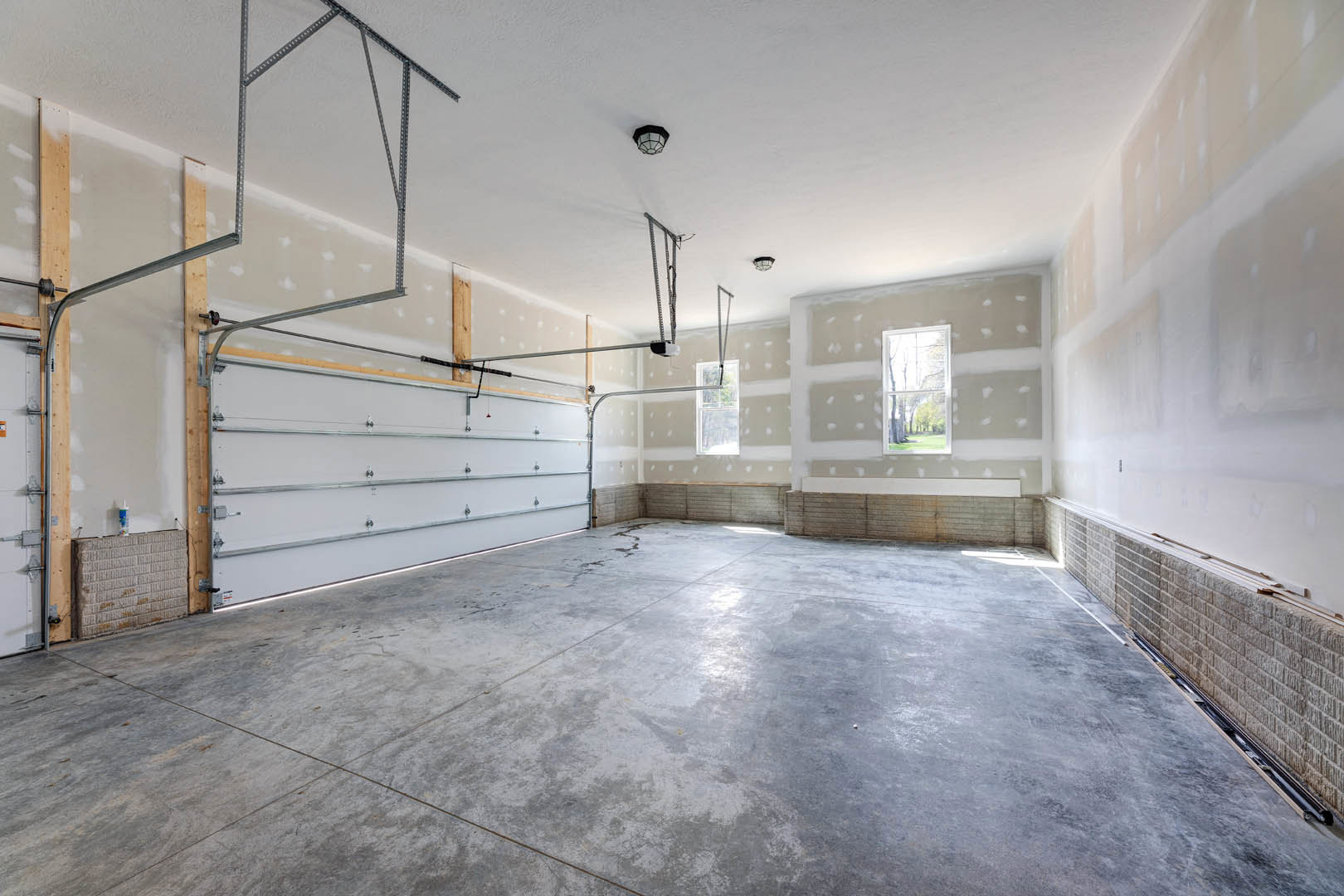 Spacious empty garage with smooth white plaster walls, white ceiling, concrete floor, metal pipes along one wall, large garage door, and window overlooking trees.