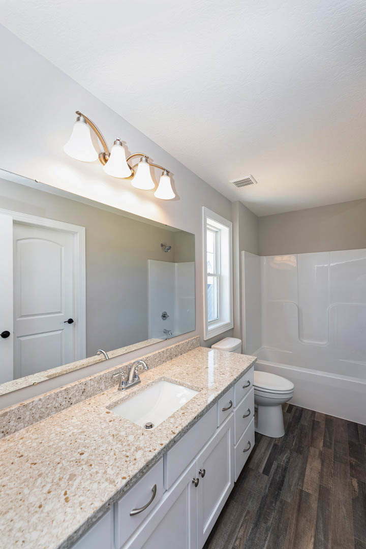 Bathroom with marble countertop, undermount white sink, chrome faucet, wall-mounted mirror, toilet with closed lid, white-framed window, light fixture, and tiled floor
