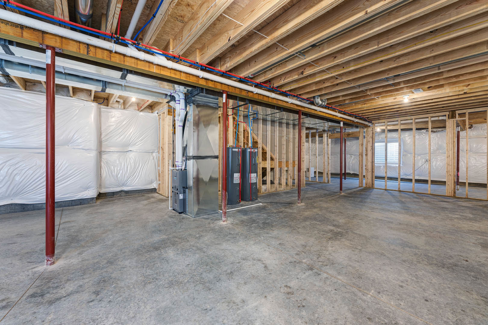 Spacious basement with exposed wood beams, concrete floor, black tanks, visible steel pipes, and large white plastic bags attached to wall