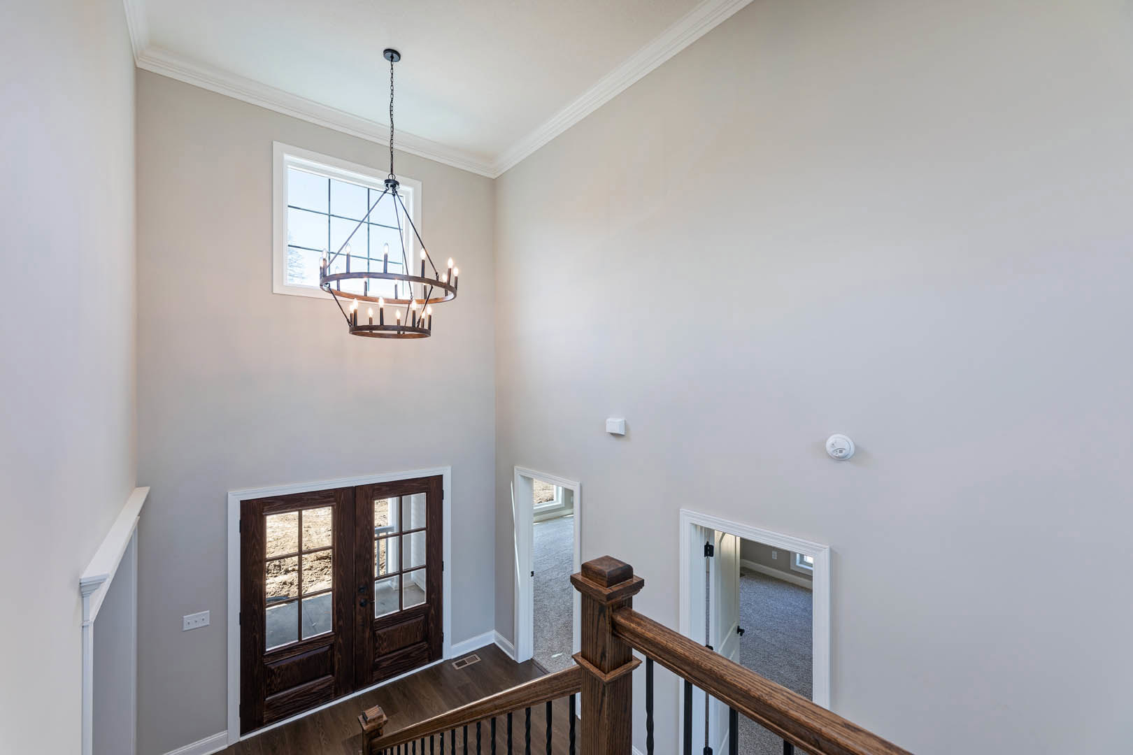 Chandelier with multiple lights hanging from a white ceiling, double glass-paneled doors, wooden floor with vent, open doorway leading to another room, staircase with white