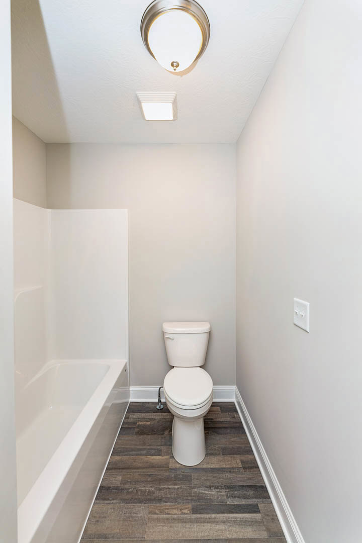 White toilet with closed lid beside a tiled wall, recessed ceiling light above, partial view of white bathtub in foreground