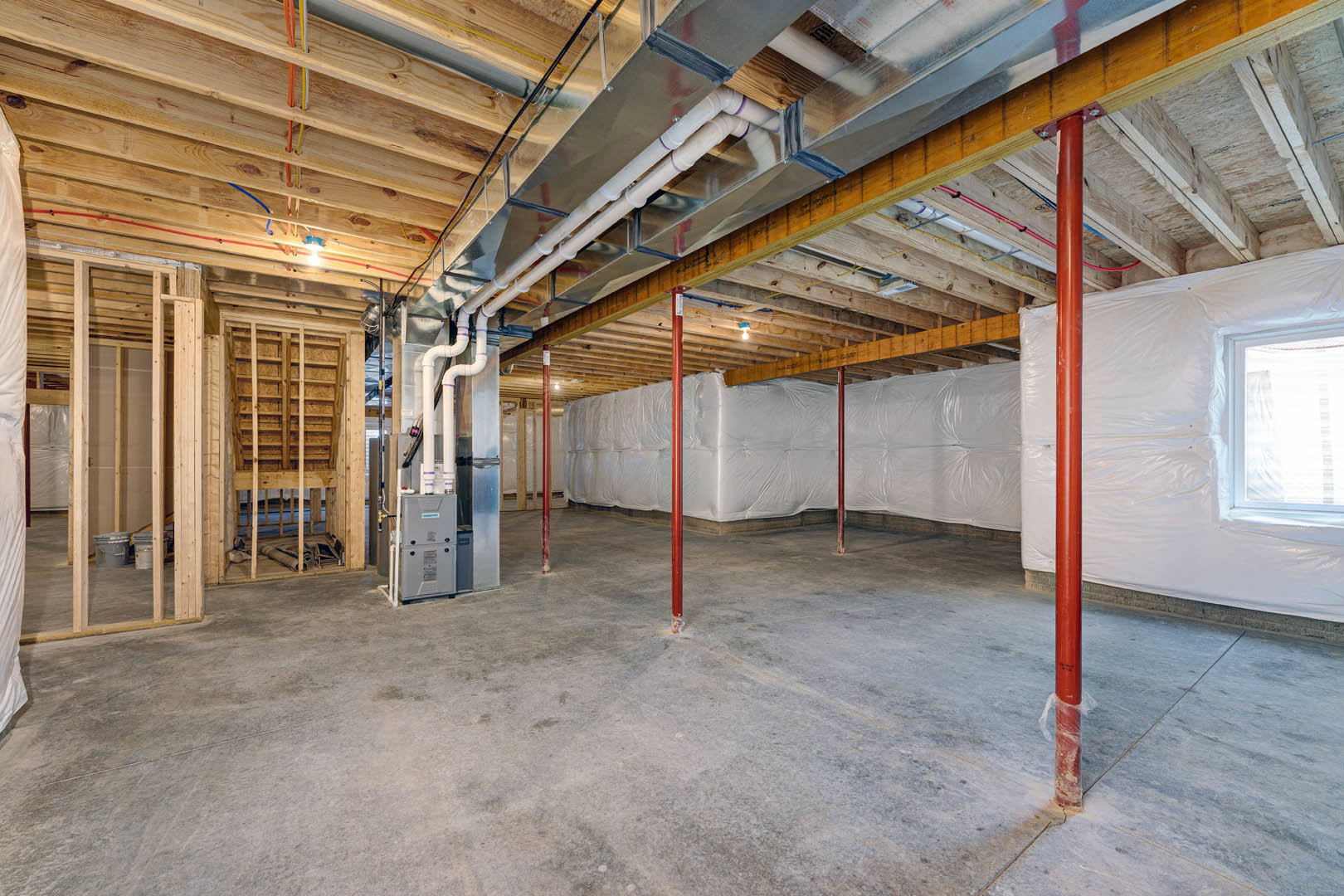 Basement room with exposed metal pipes and beams, unfinished walls, wooden staircase, grey bucket with handle, window with white curtain, and visible insulation.
