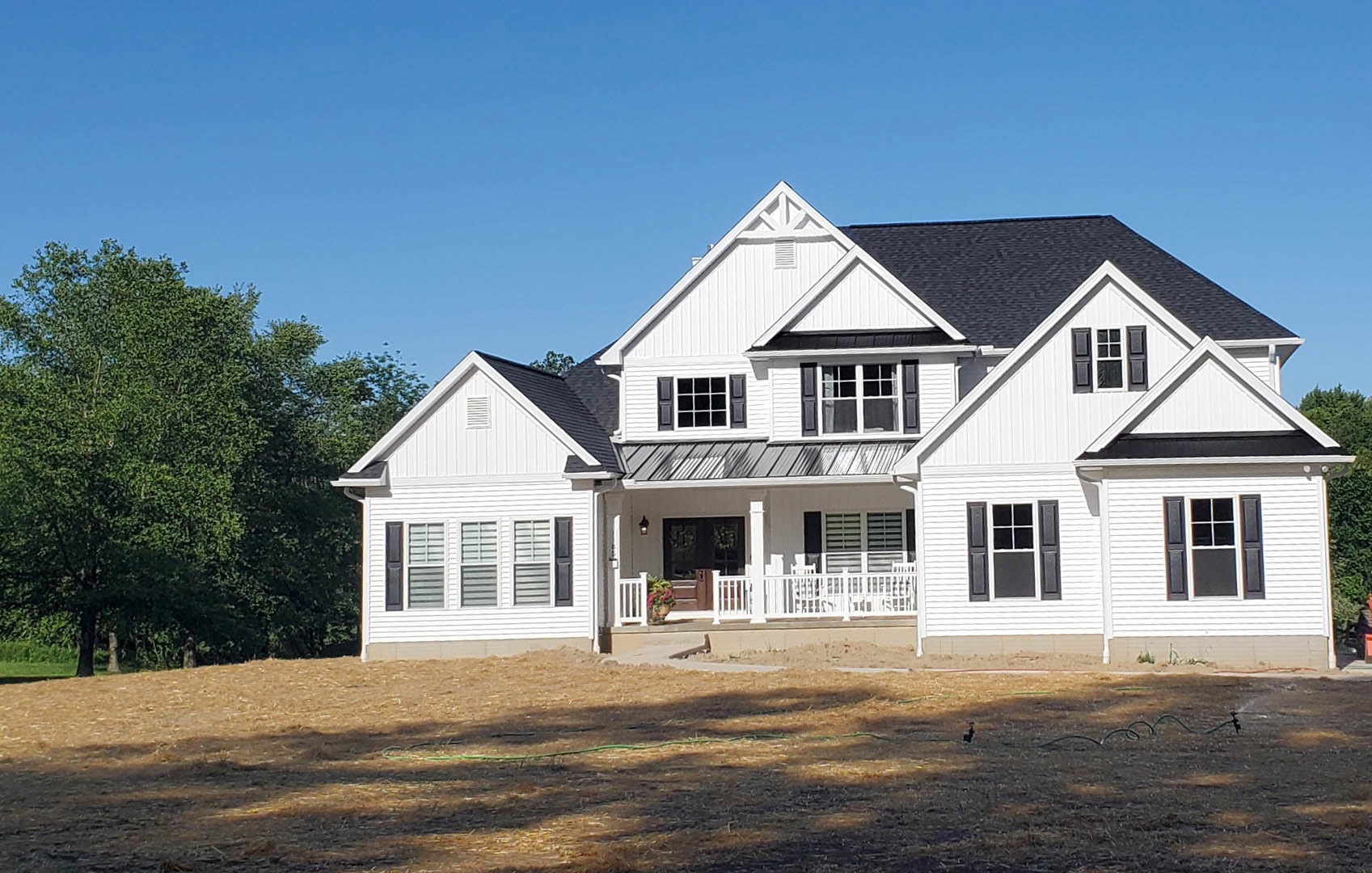 White house with black roof, expansive front porch, multi-pane windows, brown grass lawn with green hose, trees in background
