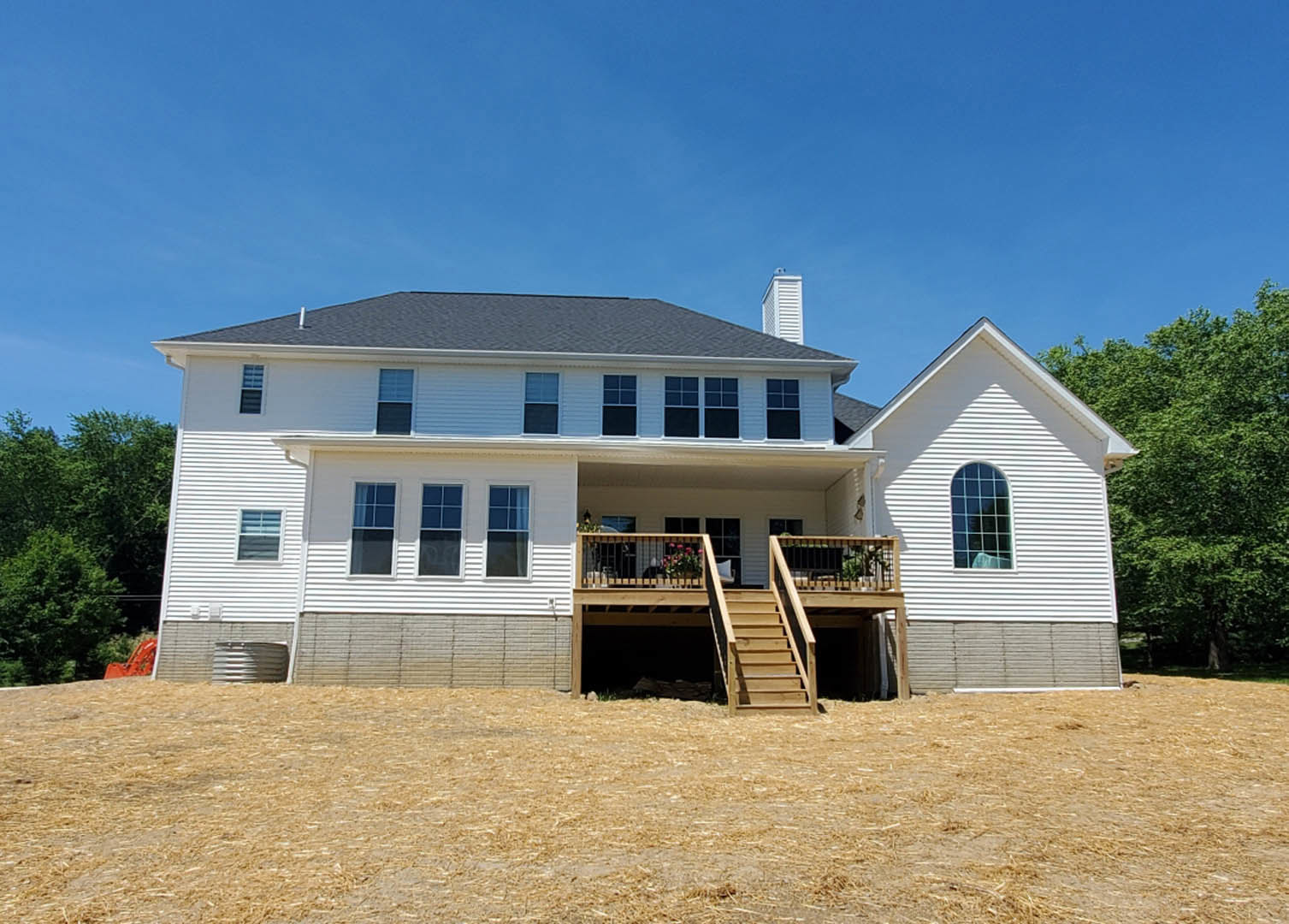 Two-story home with light siding, wooden deck and staircase, grassy yard, large windows, and mature trees in the background