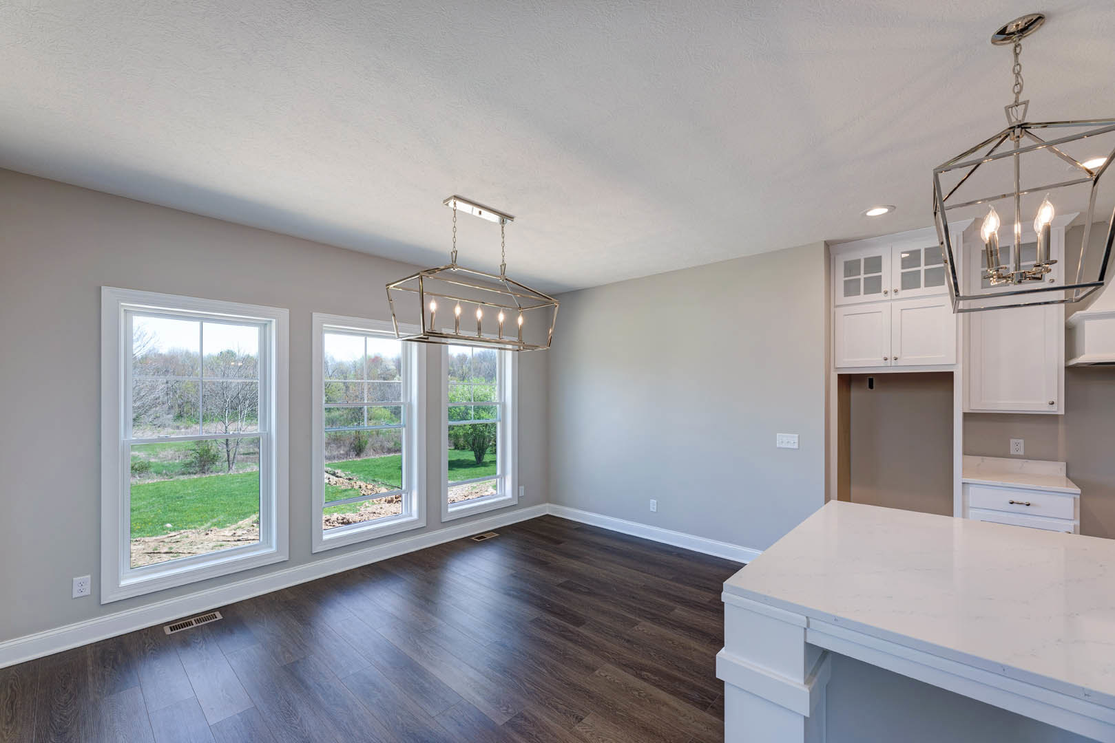 Kitchen with white quartz countertop, light wood cabinetry, hardwood flooring, large windows overlooking green lawn, modern chandelier hanging from ceiling