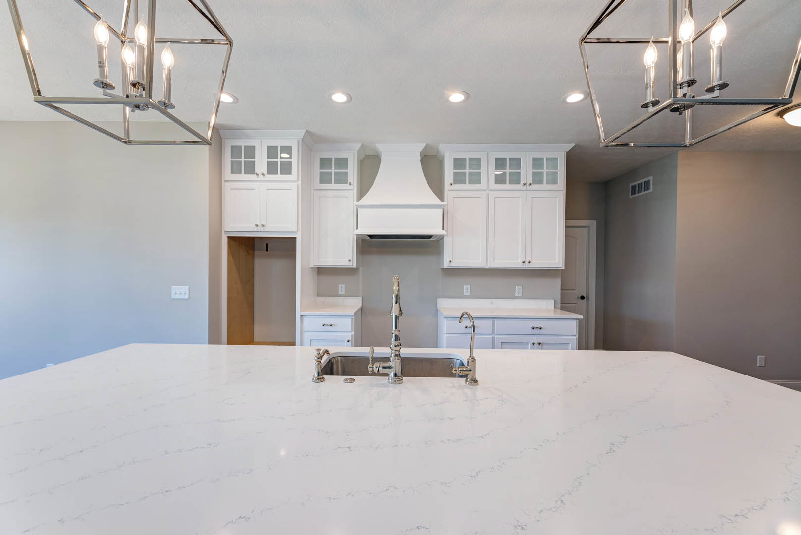 White kitchen with shaker cabinets, farmhouse sink, stainless faucet, quartz countertop, pendant lights, white vent hood, and neutral walls
