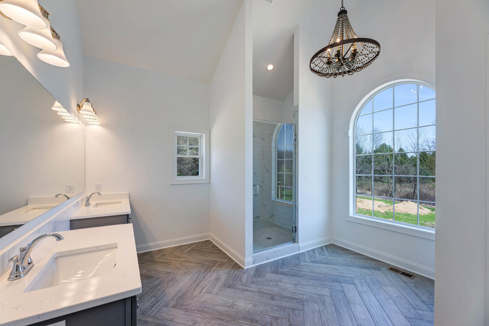 Bathroom with large multi-pane window overlooking trees, ringed bead chandelier, tiled floor and walls, modern sink with chrome faucet, mirror, and shower enclosure.