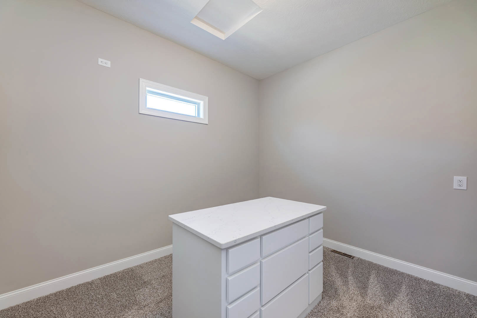 White countertop with built-in drawers beneath a window, white walls, electrical outlet, and light wood flooring in a bathroom.