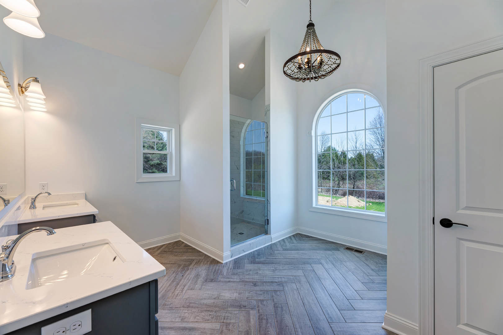 Bathroom with multi-light chandelier, wood flooring with white trim, white sink and faucet, large multi-pane window overlooking trees.