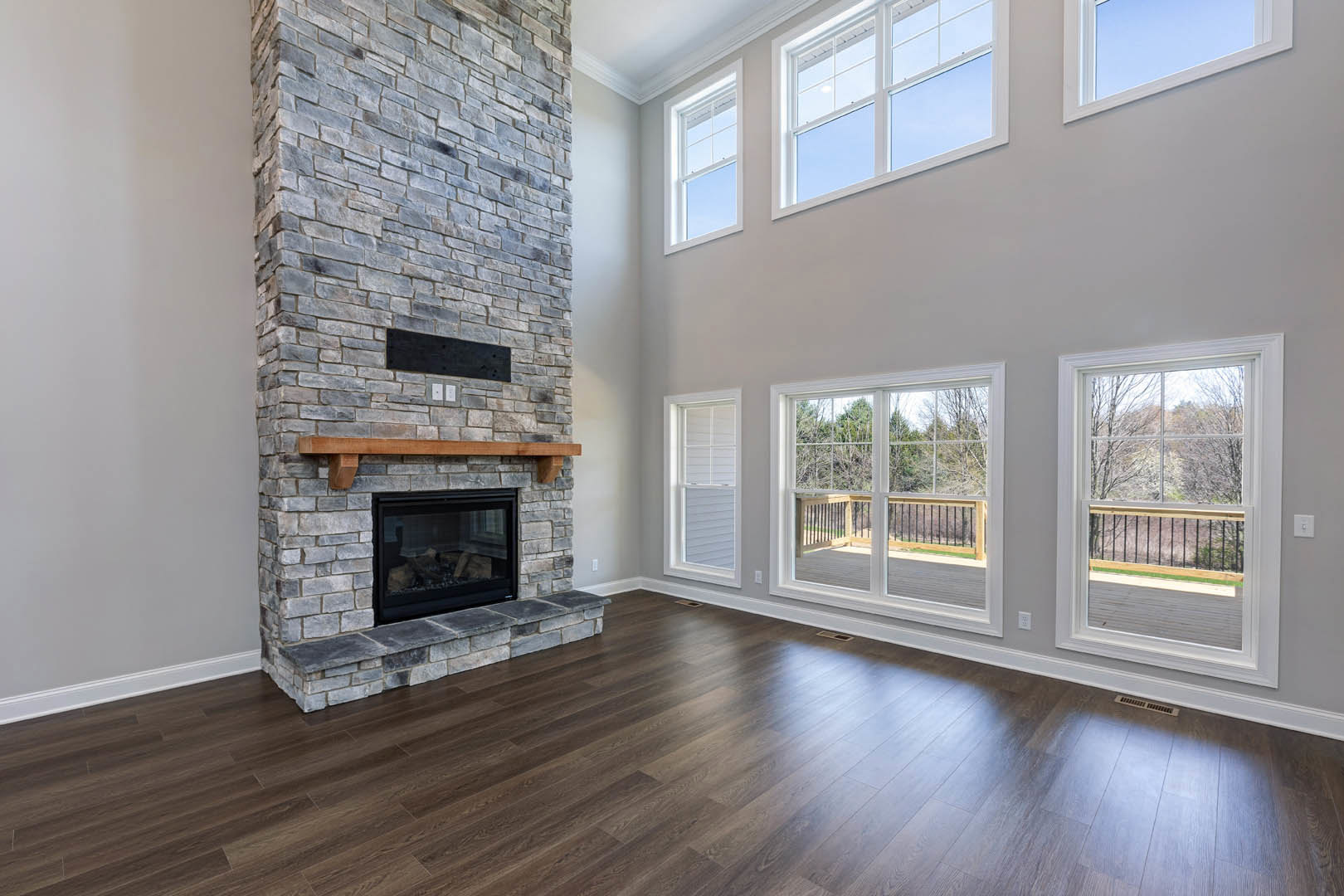 Living room with hardwood floors, brick fireplace featuring a glass door, large windows overlooking trees, and exposed wood beam accents.
