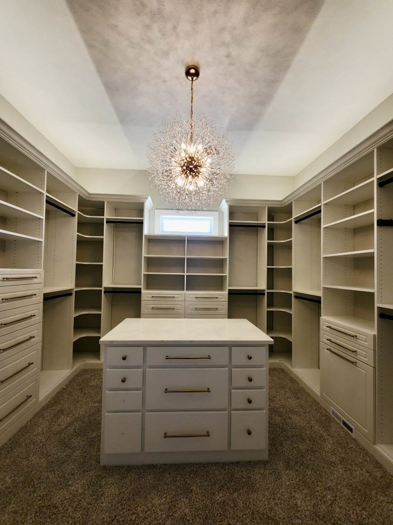 Spacious white walk-in closet featuring built-in shelves, drawers with white countertop, and a crystal chandelier hanging from the ceiling