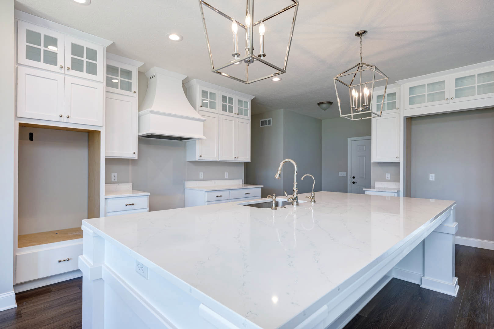 White kitchen with shaker cabinets, spacious island featuring white countertop and silver faucet, three-light pendant fixture above, white vent hood, tile backsplash, and hardwood