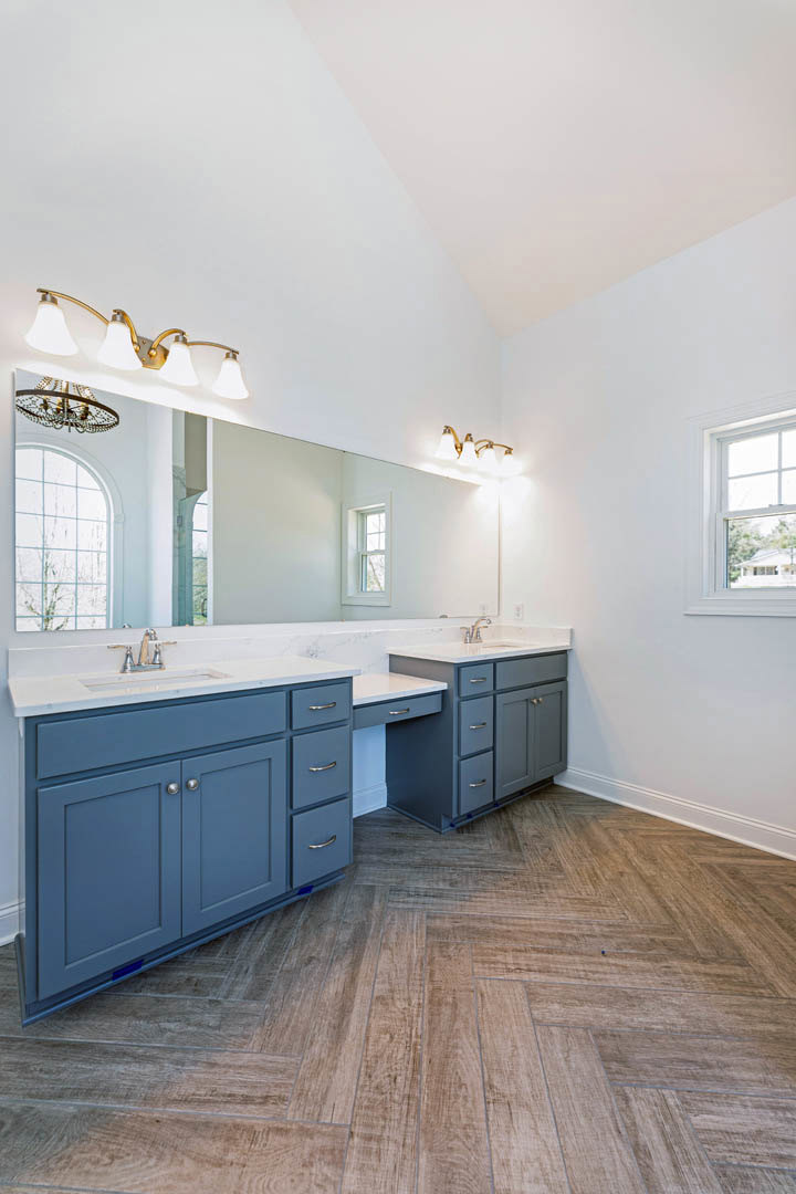 Bathroom with double sinks, wide mirror above blue vanity, white framed window, light fixture with three bulbs, tile floor, and view of a tree through the window