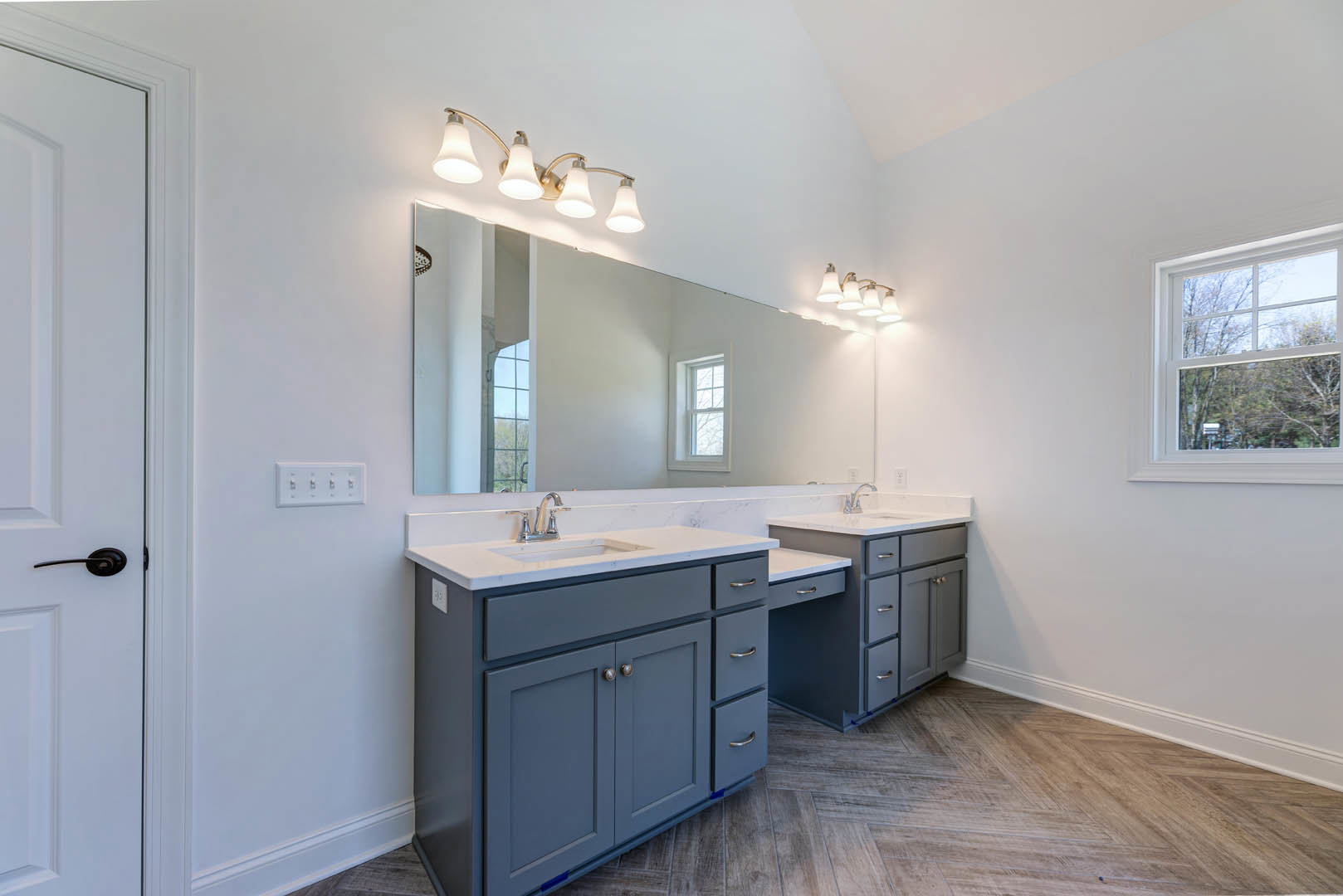 Bathroom with double sinks, wide framed mirror, white cabinetry and drawers, quartz countertop, three-light fixture above, window showing trees outside, partial view of white door.
