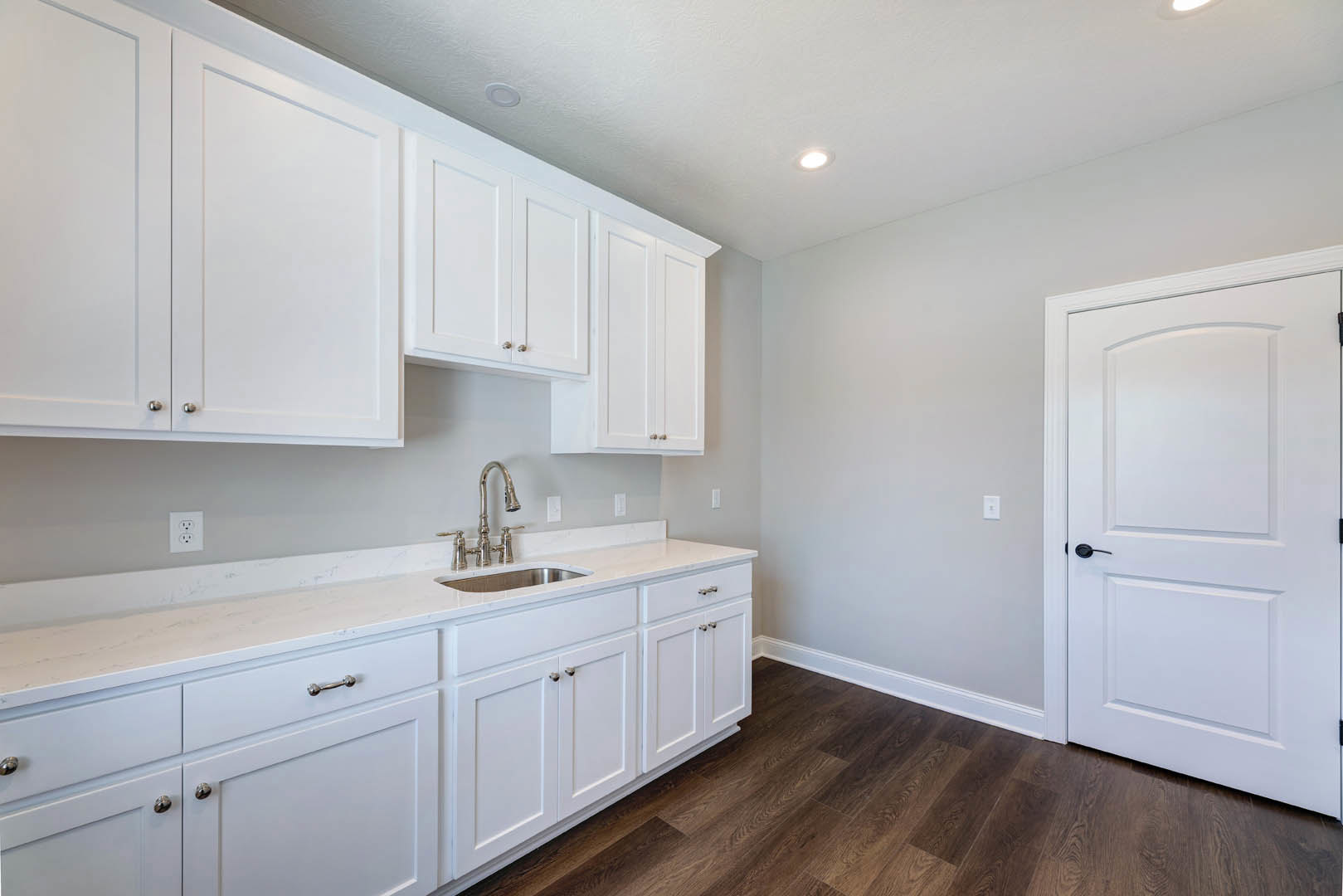 White kitchen cabinets with black handles, wood flooring, white baseboards, stainless steel faucet, and a white door with a wooden frame.
