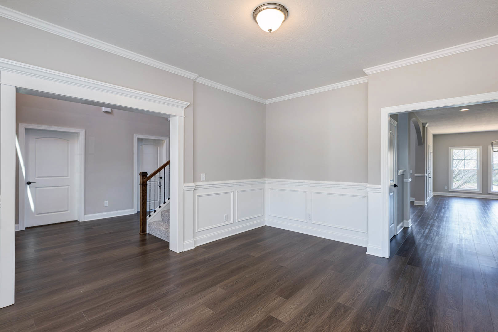 Wood flooring and white walls in a bright room, featuring a ceiling light fixture, window with white trim, close-up of a staircase, and a white door with a black handle