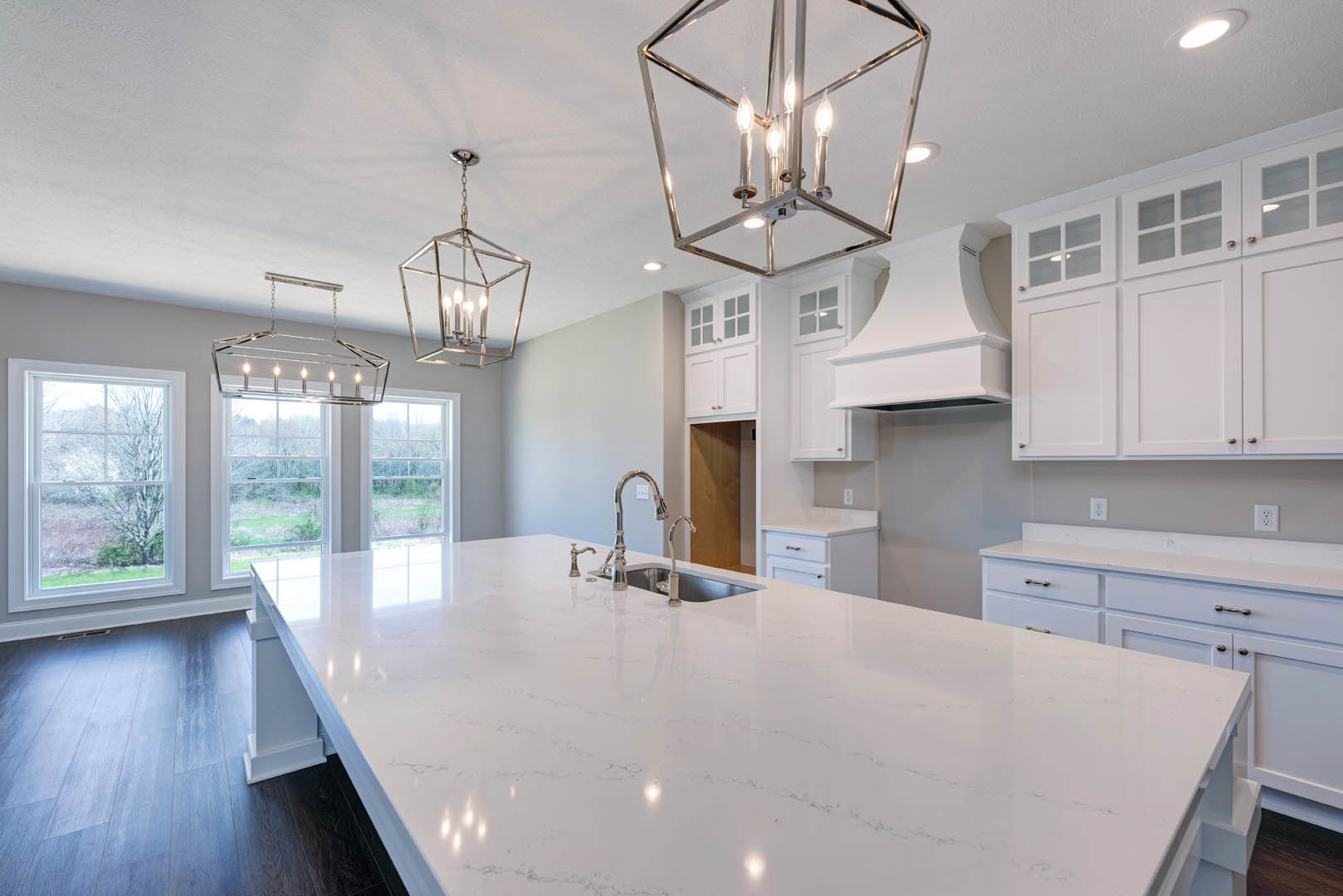 Spacious kitchen featuring a large white island with built-in sink and faucet, white cabinetry, ceiling light fixture, chandelier, and a window overlooking trees