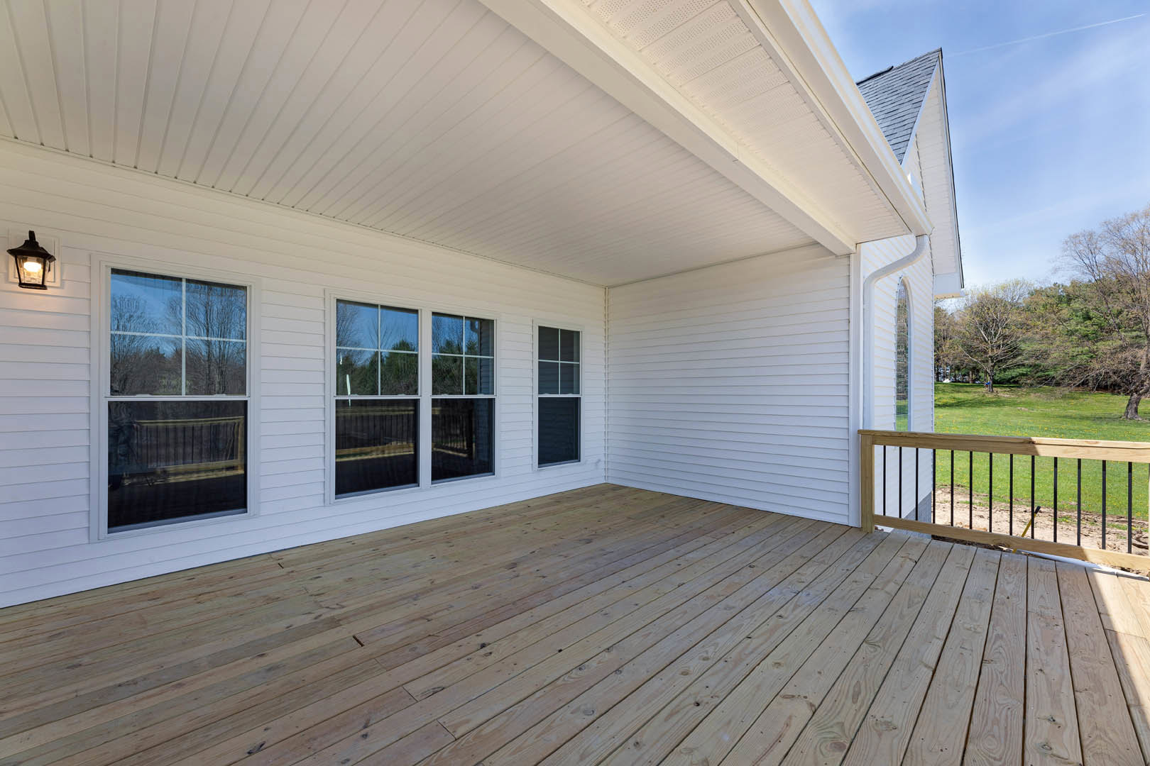 Wood deck with wooden railing, white siding, large windows reflecting trees, outdoor porch area under clear sky