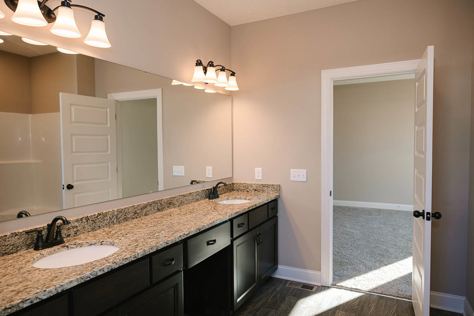 Bathroom with expansive mirror above double sinks, granite countertop, white cabinetry, chrome faucets, wall-mounted light fixture, and adjacent white door.