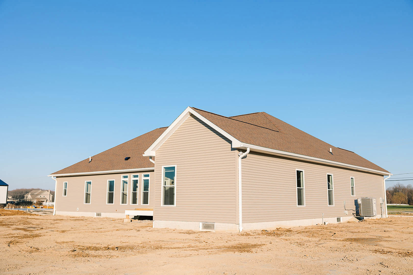 Partially built desert home with tan stucco exterior, multiple white-framed windows, large white air conditioning unit, unfinished dirt yard, and flat roof under clear blue sky