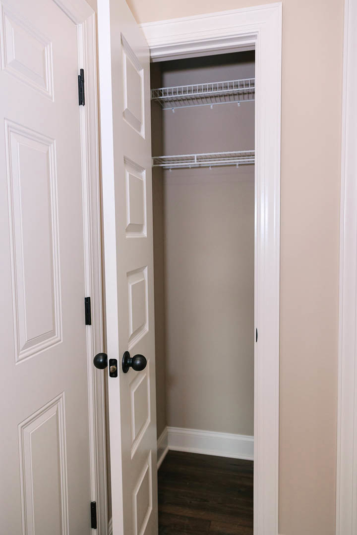 Open white closet door with brushed metal knob, dark wood flooring, white trim, built-in shelf, and wall sconce.