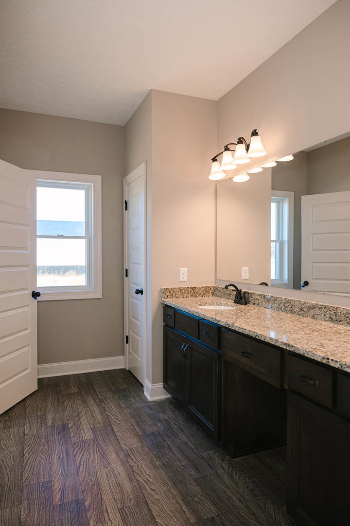 Bathroom with marble countertop, rectangular mirror above sink, white-framed window, white door with black handle, wall-mounted light fixtures, dark wood flooring with white trim
