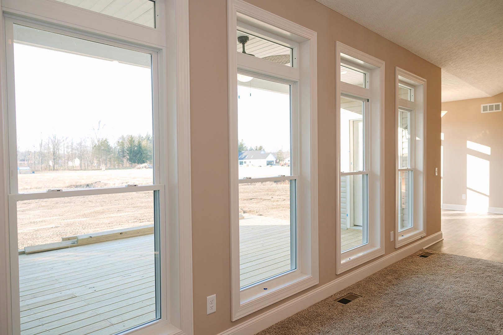 Spacious living area with multiple large windows, carpeted floor, square ottoman, white door letting in natural light, views of grassy field and trees outside