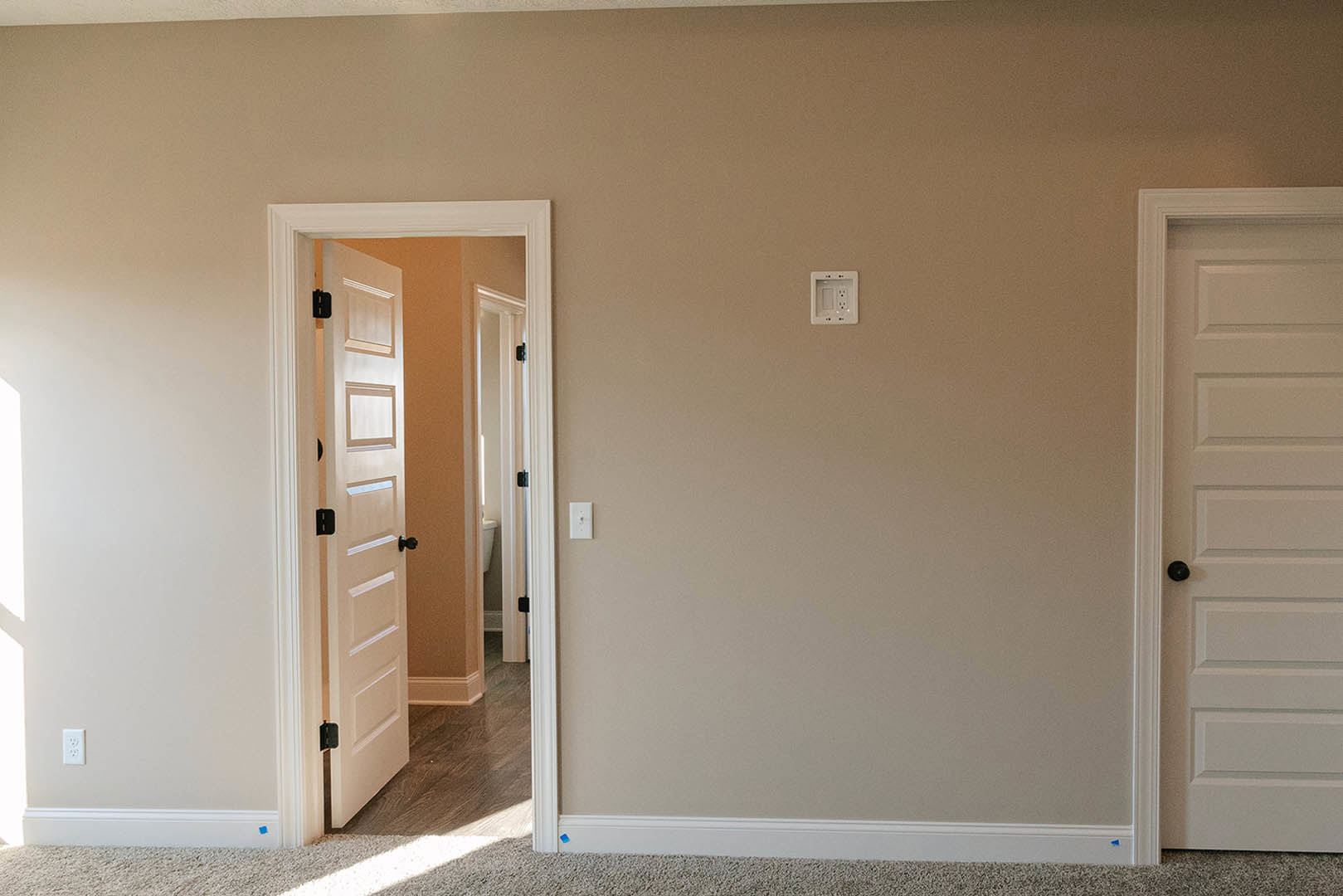 White paneled door with black knob partially open to reveal light-colored room, white electrical outlet on adjacent wall, hardwood flooring visible