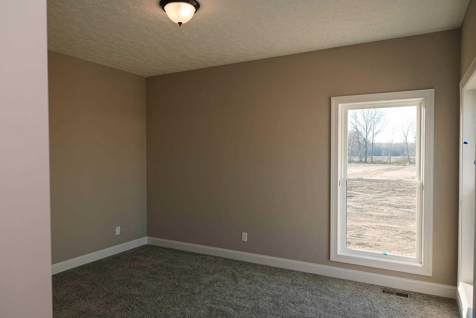 Neutral-toned carpeted floor, white plaster walls, large window with view of grassy field and trees, recessed ceiling light fixture, simple crown molding