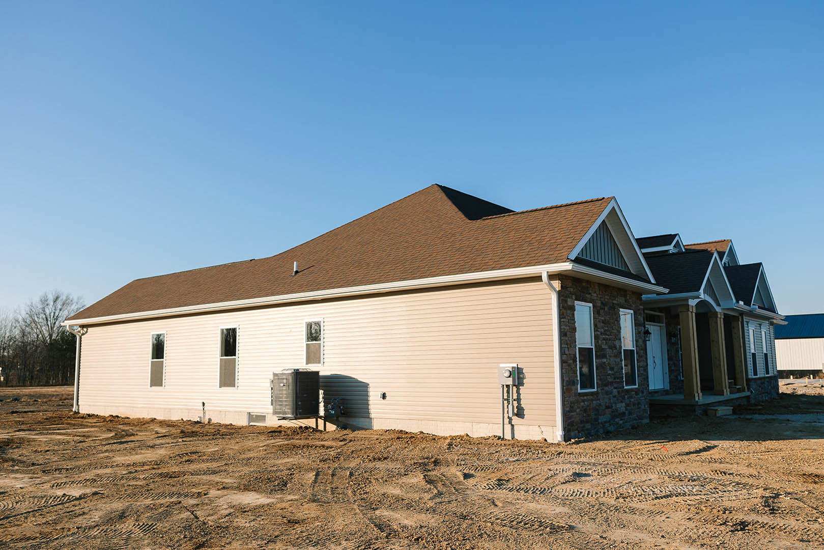 Tan-roofed house under construction with exposed dirt yard, white siding, large windows, and metal utility box mounted on exterior wall, surrounded by trees under clear sky