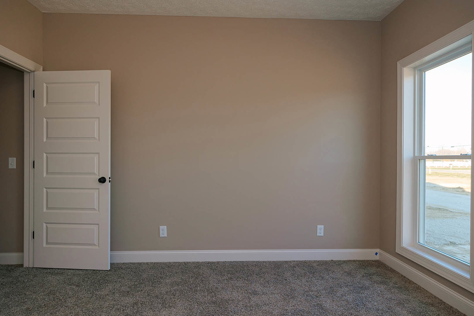 Neutral-toned carpeted room with white paneled door, large window overlooking snowy field, white plaster walls, and simple window blinds