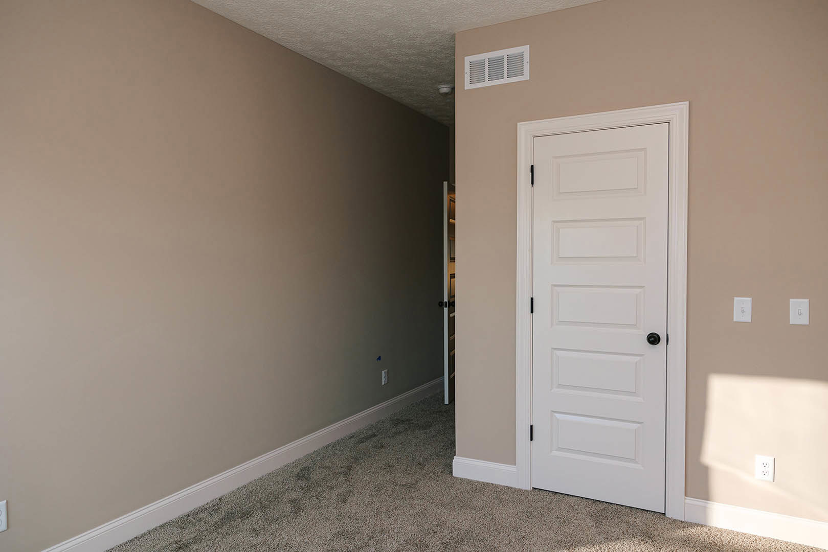 White paneled door with black knob set in a light gray plaster wall, white baseboard trim, beige carpet flooring, rectangular wall vent, and electrical outlet visible nearby