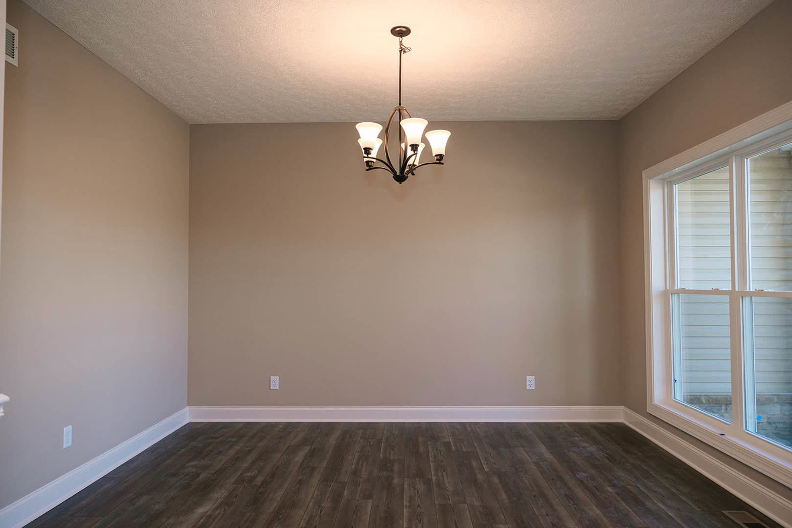 Chandelier hanging from white plaster ceiling above hardwood floor with white baseboards, large window letting in natural light