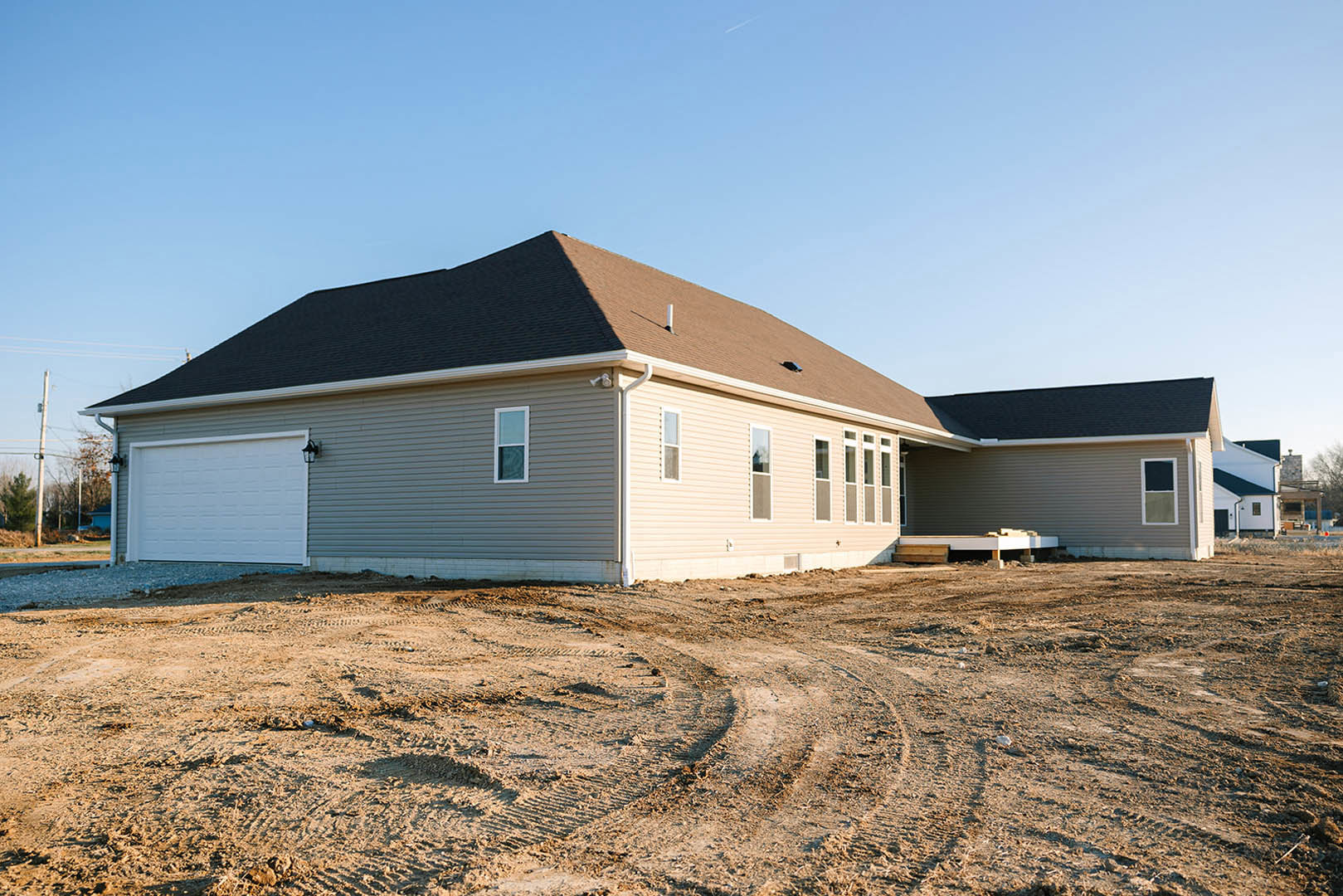 Partially built house with white siding, black roof, and white garage door, surrounded by dirt with tire tracks under a clear blue sky