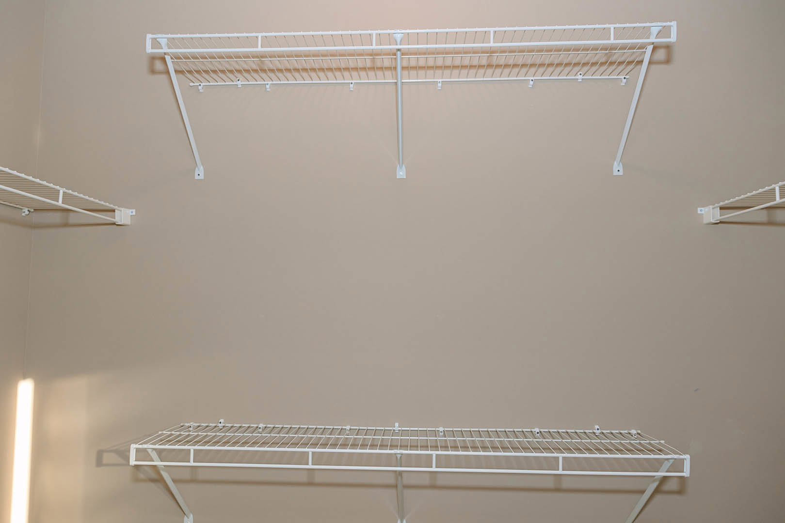 White metal shelving with pegs mounted on a white wall, illuminated by natural light from a nearby window, with a white pipe running along the wall.