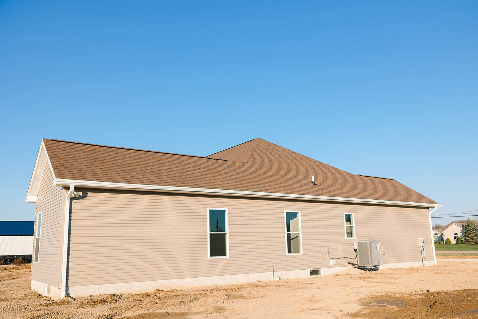 Partially constructed house with white-framed windows, gray siding, and roof under installation; heat pump unit visible near foundation; tree and blue sky in background