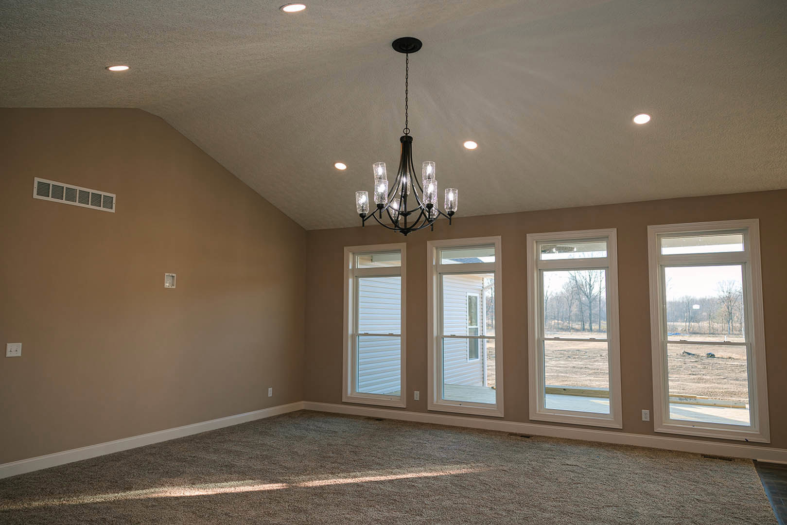 Chandelier hanging from white plaster ceiling, large windows with views of trees and open field, light-colored walls with decorative molding, hardwood flooring, wall vent visible.