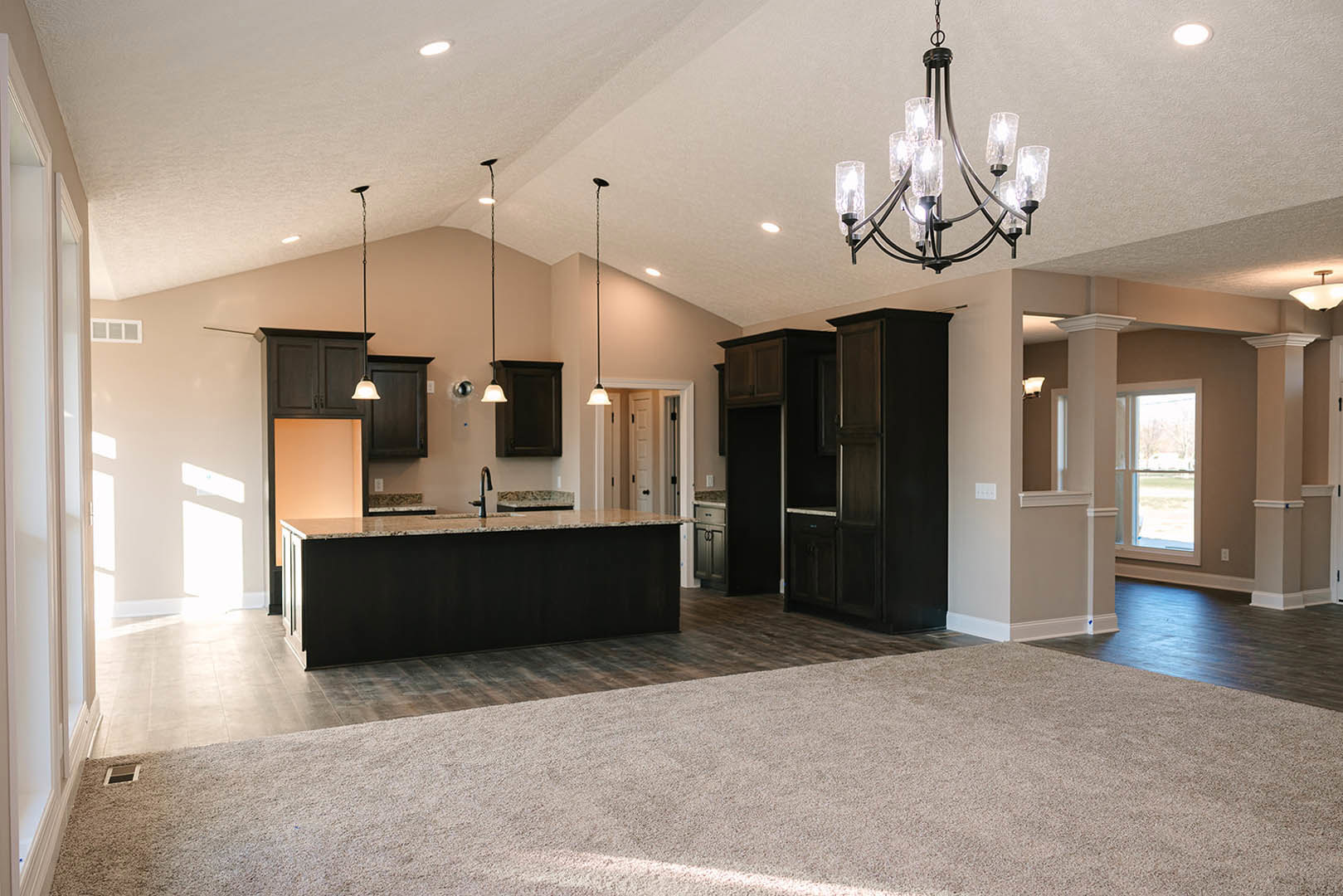 Open-concept kitchen and living room featuring black granite countertops, white cabinetry, a carpeted floor, clear glass chandelier, and large window overlooking grassy field