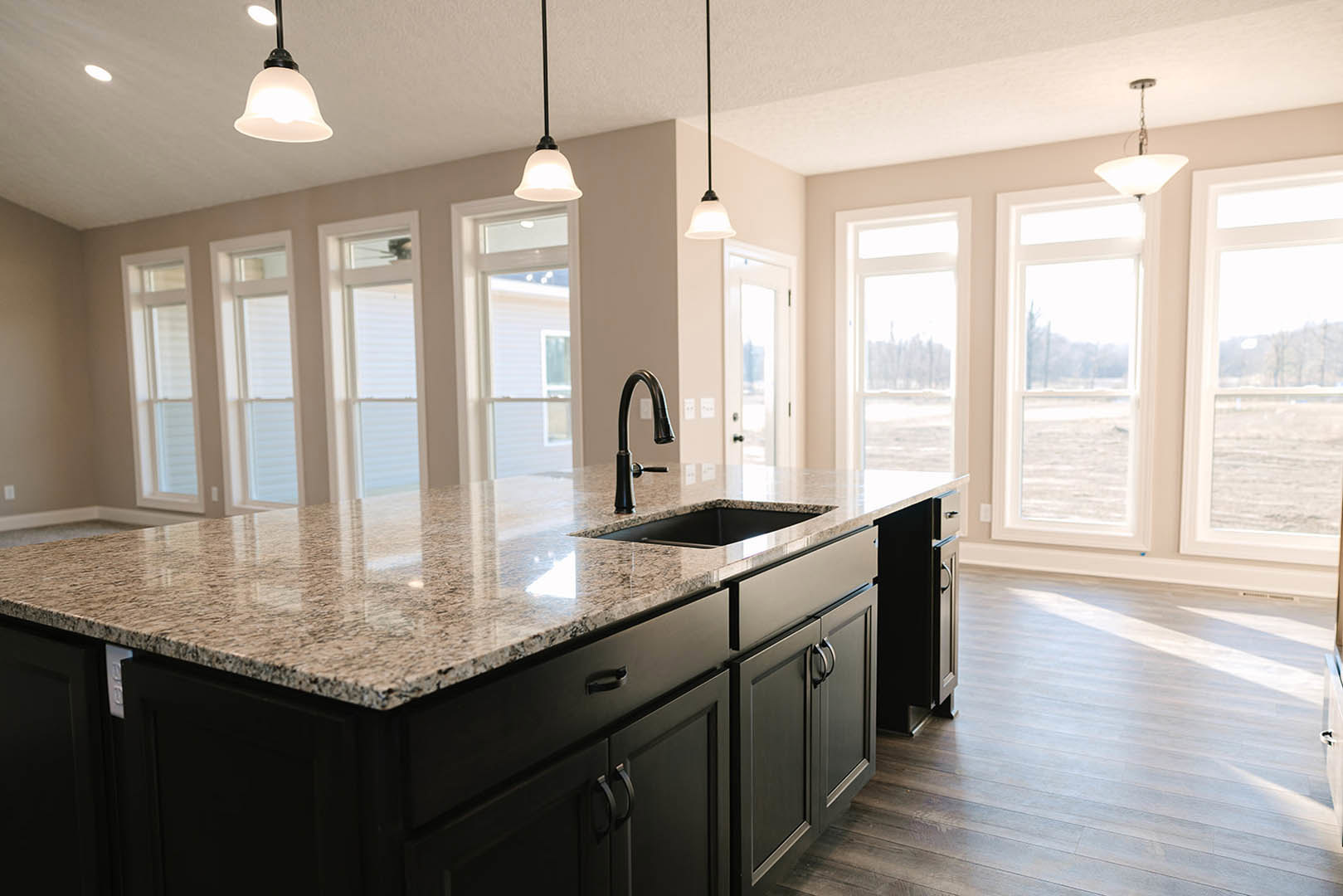 Spacious kitchen featuring a large island with black countertop, built-in sink and modern faucet, white tile backsplash, light wood cabinetry, pendant light fixtures, and a window