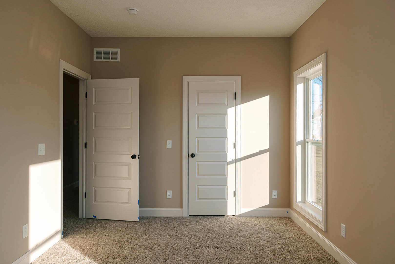 White-walled room with two paneled doors featuring black handles, single window with white trim, beige carpet flooring, and wall vent