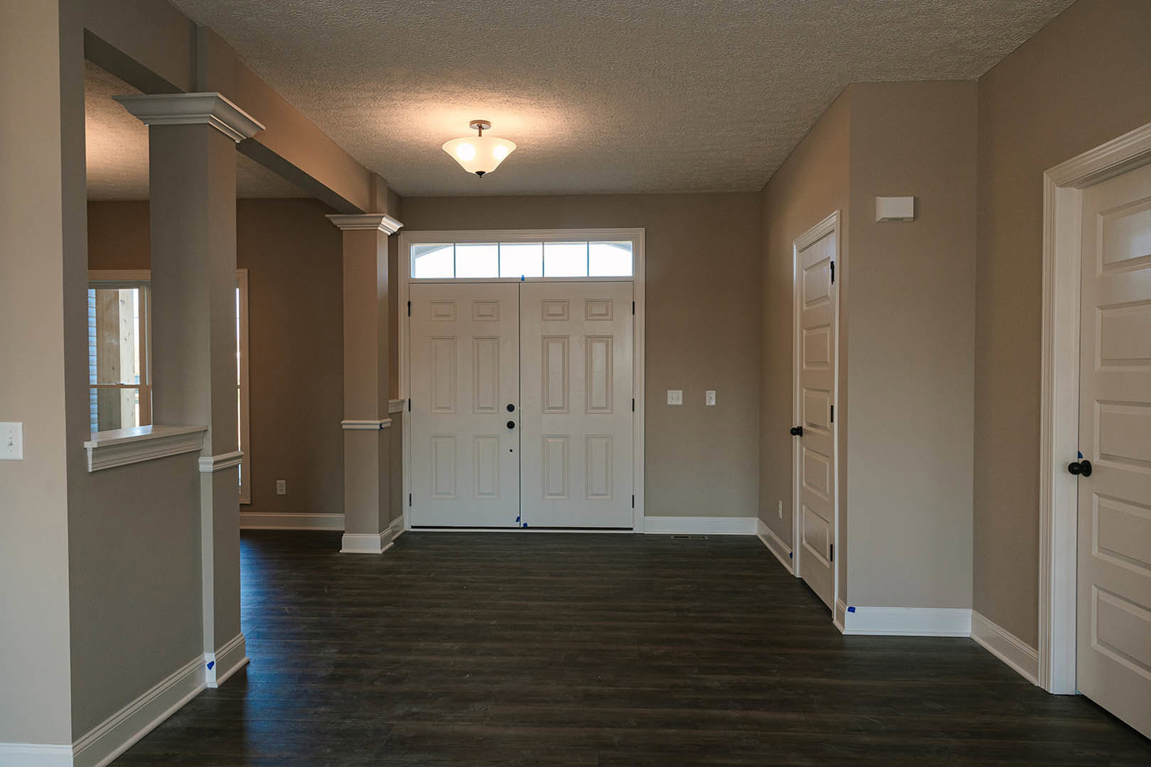 Hallway with dark wood laminate flooring, white trim, multiple white doors including double doors with glass window, three-light ceiling fixture, and a window featuring a blue