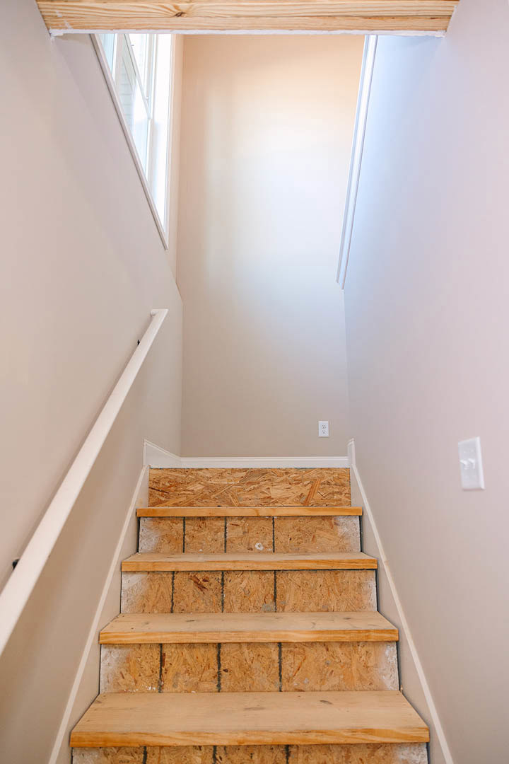 Modern staircase with plywood side panels, white plaster walls, minimalist handrail, and built-in wooden shelf near a window